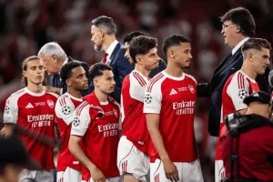 Arsenal players in red and white kits lined up on the pitch before a match, with coaches nearby.