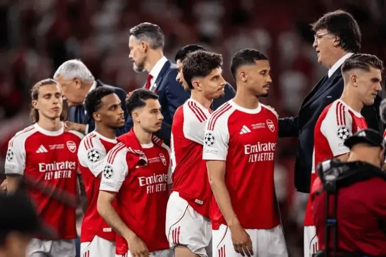 Arsenal players in red and white kits lined up on the pitch before a match, with coaches nearby.