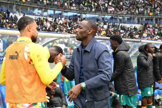 Handshake on the soccer sideline between a man in a dark suit and a player in a yellow training vest, with teammates and a crowded stadium in the background.