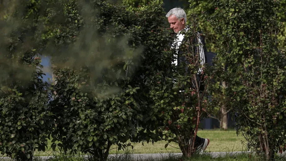 Older man in a striped blazer walks along a path behind dense shrubbery in a park.