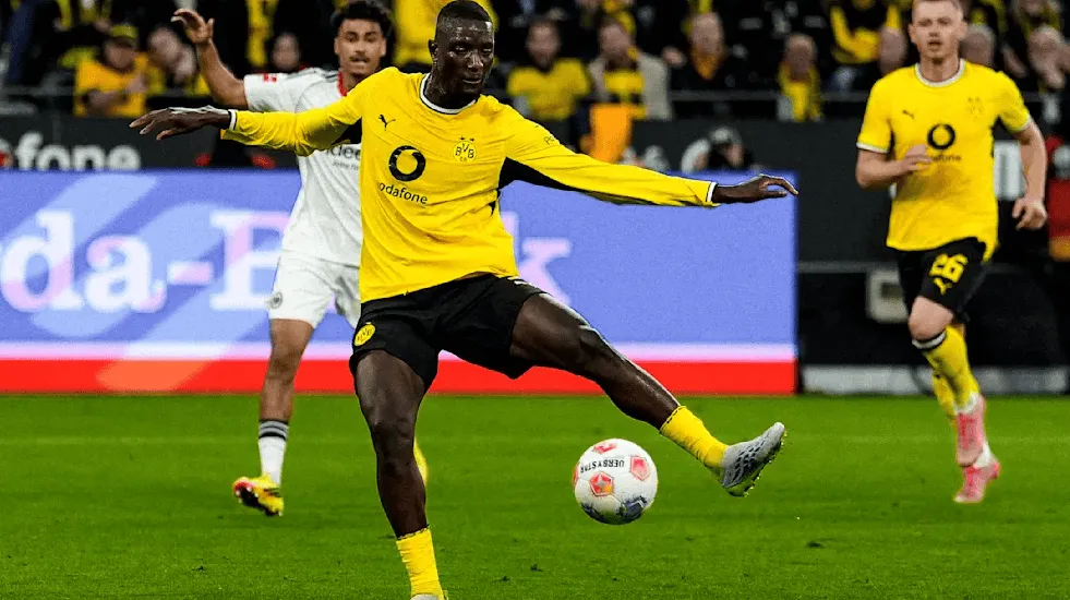 Soccer player in a yellow Borussia Dortmund jersey kicking a ball on a green field during a match, with teammates and fans in the background.