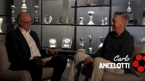 Two men sit on couches in a trophy-filled showroom, mid-conversation; glass display shelves behind them and on-screen text reads 'Carlo Ancelotti'.