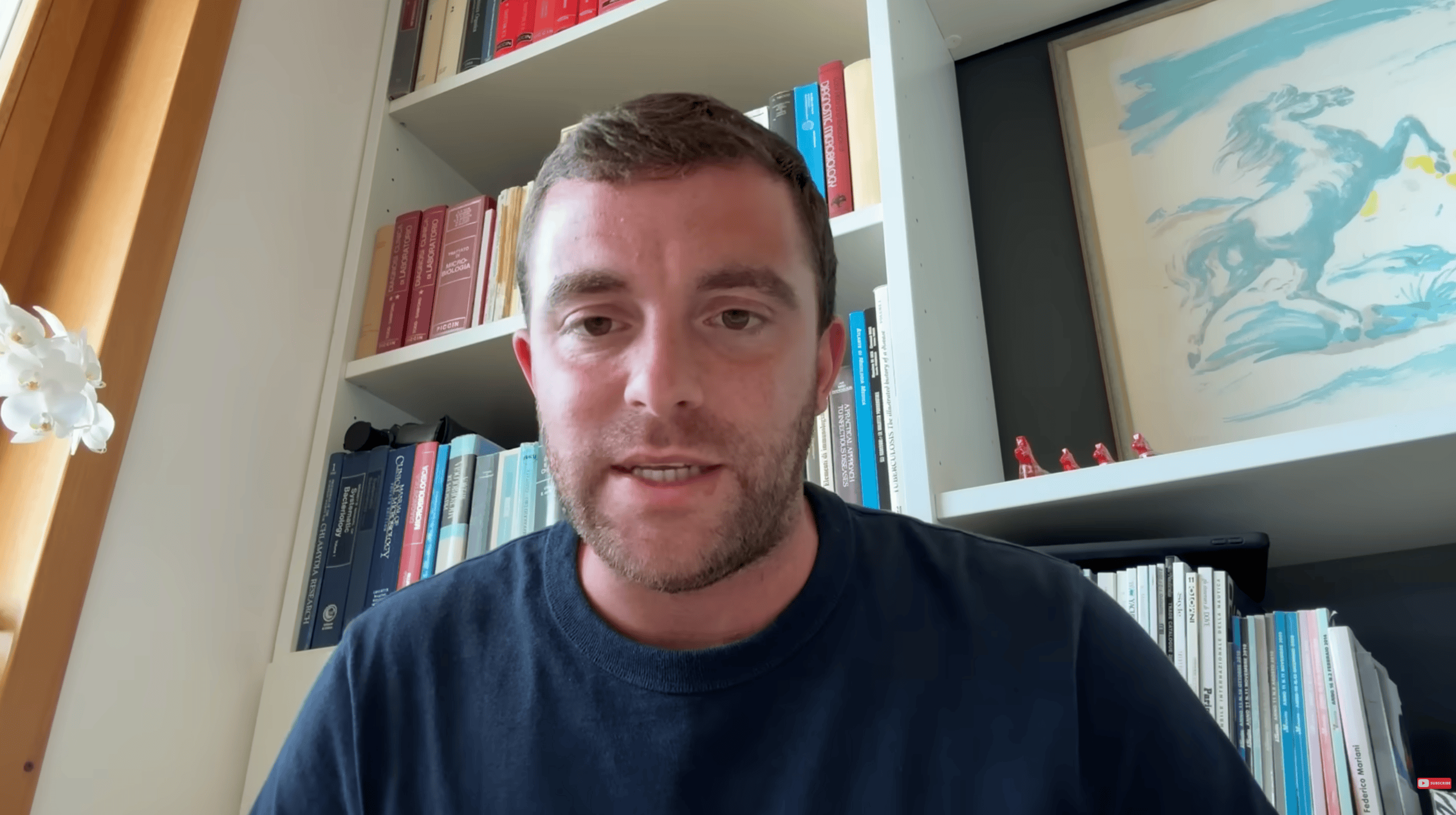 Close-up of a man with short brown hair and stubble speaking to the camera in a home office with a bookshelf behind him.