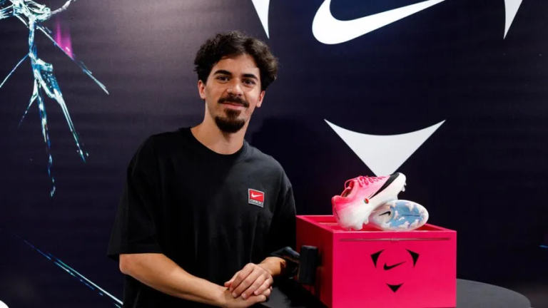 Young man in a black t-shirt poses beside a pink Nike display box with two sneakers on top.