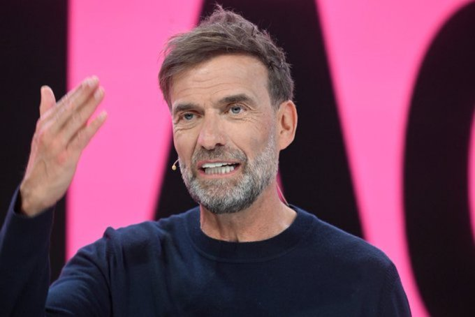 Man with gray beard speaks on stage, gesturing with his right hand, wearing a dark sweater, pink backdrop behind him.