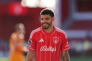 Male footballer in a red striped jersey smiling on the pitch, sponsor logos visible (Bally's) and Adidas stripes on shoulders, during a match/event.