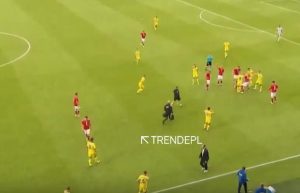 Soccer match on a green field with red and yellow team players; officials and substitutes near the center, with a white on-screen text label 'TRENDEPL'