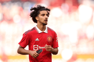 Manchester United player in a red jersey running on the field, holding shin pads.