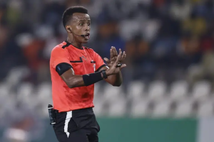 Soccer referee in an orange uniform with a headset, signaling with hands during a match