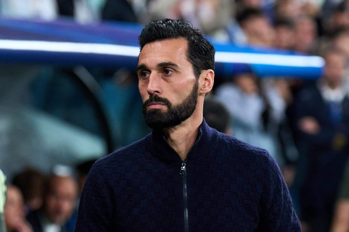 Bearded man with dark hair in a navy zip-up jacket standing in a stadium crowd, looking to the left.