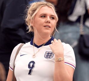 Woman wearing an England football jersey (number 9), with blonde hair, gold watch, and heart pendant necklace, looking to the side.