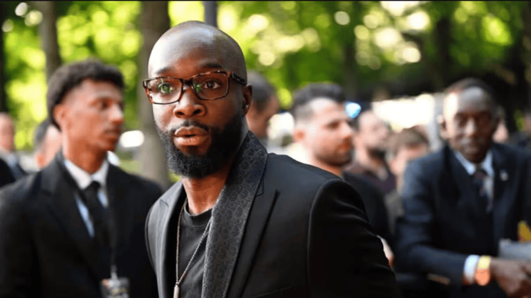 Black man with a shaved head and beard, wearing glasses and a black suit, looking toward the camera at an outdoor event.