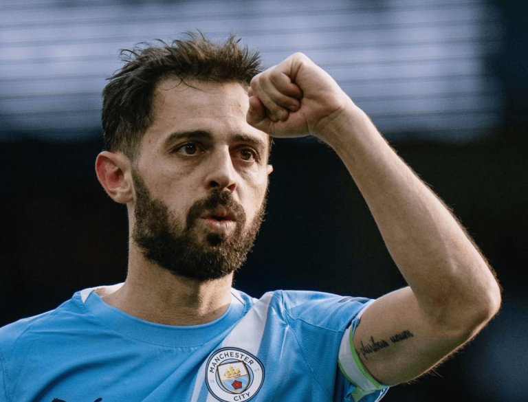 Male soccer player with a beard in a light blue Manchester City jersey raises his right fist in celebration on the field.