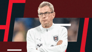Male football coach in a white training jacket with crest, arms crossed, looking serious amid a red and dark abstract background.
