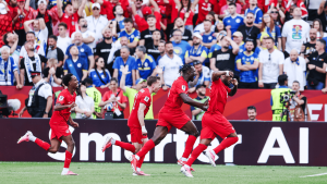 Soccer players in red uniforms celebrate a goal on the field with a cheering crowd behind them at the stadium.