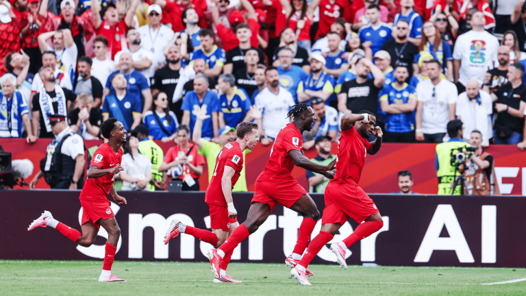 Soccer players in red uniforms celebrate a goal on the field with a cheering crowd behind them at the stadium.