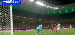 Soccer match near the goal as a blue-clad goalkeeper reaches for the ball while red and white players contest it under bright stadium lights.