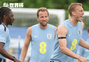 Three England footballers in light blue training shirts on a practice field, smiling and talking near a goal with a World Cup logo in the corner.